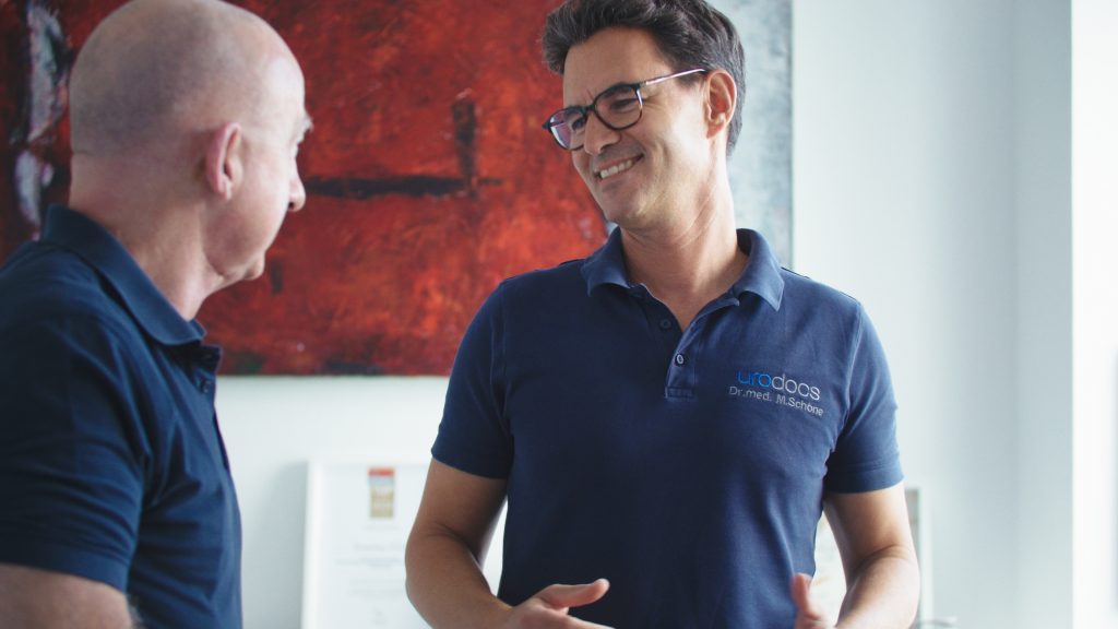 Two men in blue polo shirts talk in a bright room; one wears glasses and smiles, with a red abstract painting on the wall behind them.