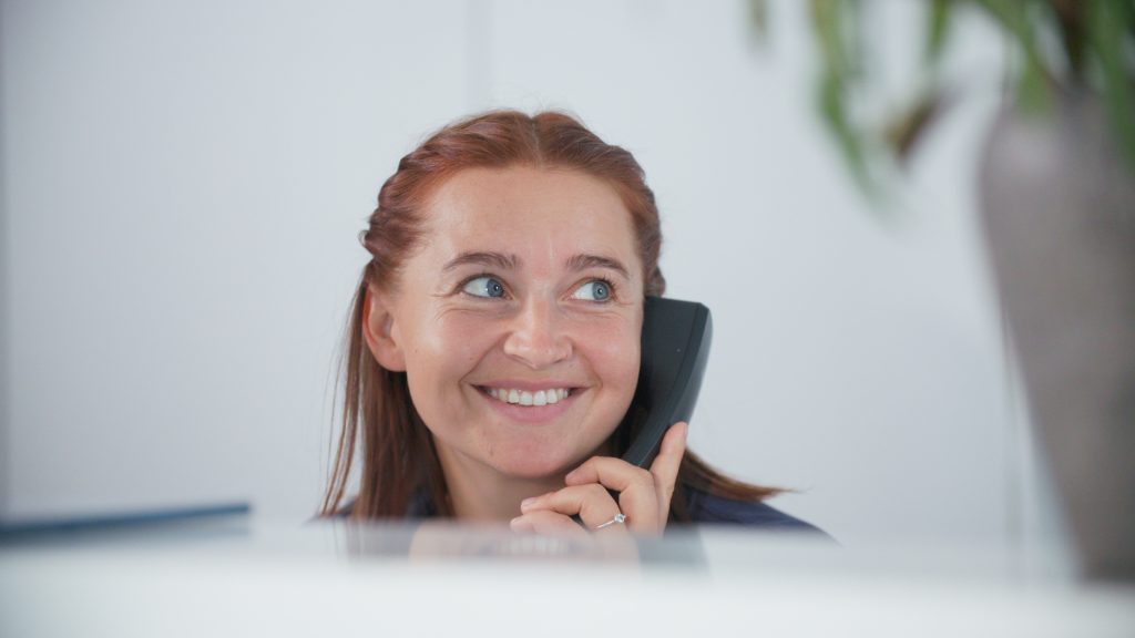 Smiling woman with red hair wearing a headset, looking off to the side in a bright office.